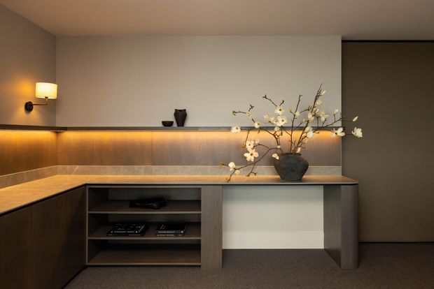 A warm, moody built-in living room cabinetry and media console. Dark timber-toned joinery runs along two walls, with open shelving, a subtle recessed display area, a pale stone or tiled benchtop, and concealed amber LED strip lighting beneath the upper ledge. Decorative branches in a dark vase sit on the counter, while a wall sconce provides additional soft illumination. A dark doorway or wall opening is visible to the right, and carpet covers the floor.