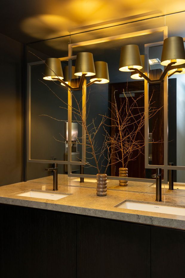 A dark, sophisticated double-vanity bathroom with a long pale stone countertop, two integrated white basins, slim dark metal faucets, and black fixed cabinetry below. Large framed mirrors span the wall, reflecting sculptural multi-arm pendant or wall-mounted lighting and warm illuminated ceiling details. Decorative bare branches in metallic vases add an organic accent to the moody, luxurious space.