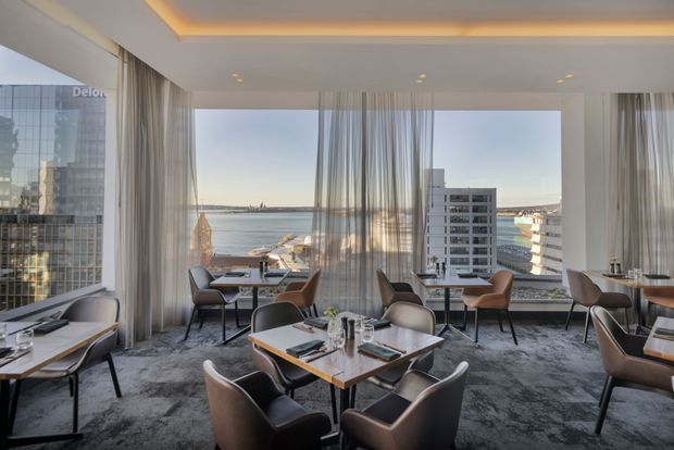 A refined high-rise dining restaurant or lounge with floor-to-ceiling glazing overlooking a harbour and city skyline. The space features multiple small dining tables, sculptural upholstered chairs in brown and charcoal tones, dark patterned carpet, sheer full-height curtains, and a recessed ceiling with warm cove lighting.