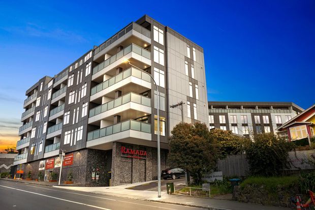 Exterior view of a contemporary multi-storey apartment or hotel building at dusk, featuring dark cladding, light panelled facades, stacked balconies with translucent glass balustrades, extensive vertical glazing, and ground-floor commercial signage. A smaller traditional house is visible beside the building, with landscaped planting along the street.