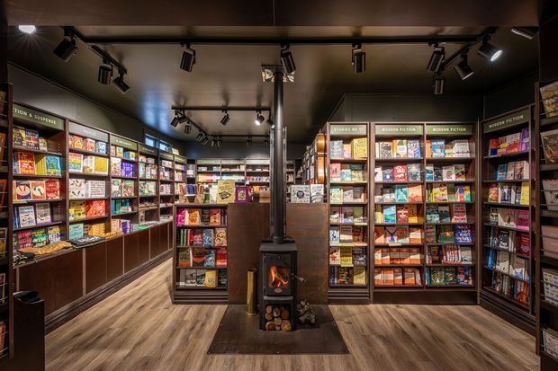 A warmly lit specialty retail interior resembling a bookstore or magazine shop, with floor-to-ceiling shelving densely stocked with books, magazines, and board games. Dark timber-toned cabinetry lines the walls, while a freestanding black wood-burning stove with visible fire occupies the centre. Track lighting is mounted across the dark ceiling, and the space has a light wood-look floor.