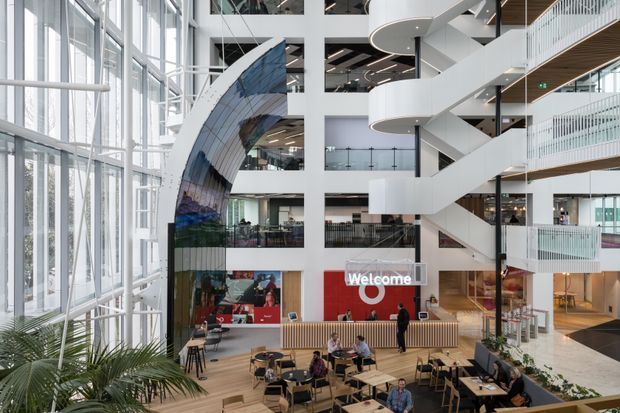 A bright, expansive multi-level commercial atrium with floor-to-ceiling glazing, open-plan seating, a reception or café-style ground-floor area, balconies and multiple circulation bridges. A prominent sculptural white open staircase and a large curved reflective feature wall define the contemporary interior.