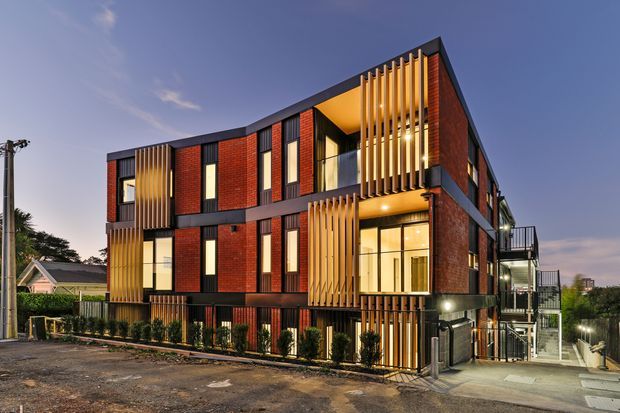 A contemporary multi-storey residential building photographed at dusk, featuring red brick walls, dark horizontal and vertical framing, warm illuminated glazing, and prominent vertical timber privacy screens across balconies and windows. The building sits on a compact urban or suburban site with an external access path and neighbouring homes visible around it.