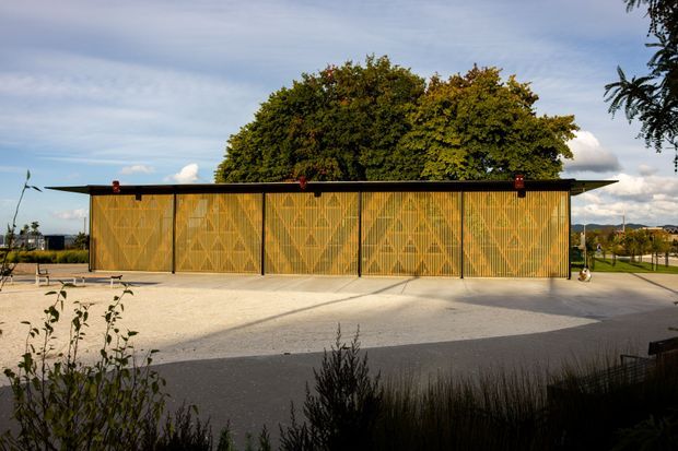 A low, elongated contemporary pavilion with a flat roof and a striking golden-brown timber screen facade composed of repeating geometric patterns. The building opens onto a broad pale paved forecourt, with mature trees behind it and landscaped grounds extending around the structure. The image is photographed from an outdoor setting in warm late-day light.