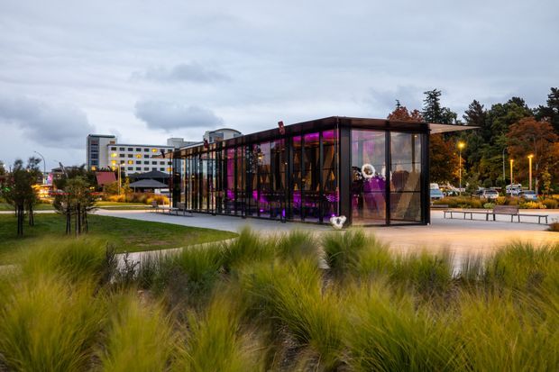 A dusk exterior view of a contemporary glass pavilion or event structure set within landscaped grounds. The long, transparent rectangular building has dark metal framing, vivid purple interior lighting, and a projecting flat roof. Ornamental grasses, paved paths, benches, lighting, trees, and a larger multi-storey building are visible in the surrounding outdoor setting.