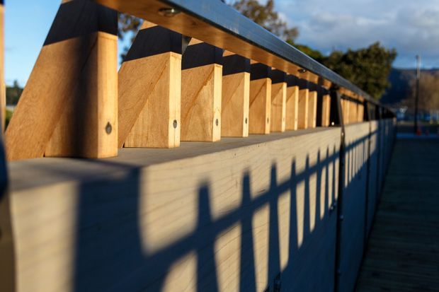 Close-up exterior architectural view of a long timber structure with repeated vertical plywood or timber fins beneath a dark metal roof edge. Strong low-angle sunlight casts rhythmic shadows across the pale wall, while a narrow timber walkway and railing extend alongside the building. The image emphasizes repetitive facade detailing, timber construction, and linear perspective.