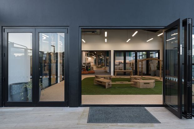 Contemporary commercial or community interior viewed from an exterior threshold, with dark metal-framed glazing and wide folding doors opening onto a bright, open-plan space. The interior features artificial grass, low timber seating and play-like built-in platforms, exposed services, and linear recessed lighting. Pale concrete or stone paving extends across the foreground.
