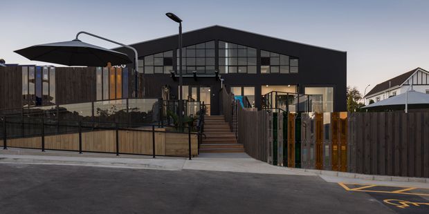 Exterior view of a contemporary dark-clad, multi-level building with a prominent glazed gable facade, rooftop or elevated terrace areas, timber fencing, outdoor umbrellas, and a street-side entrance. A neighbouring residential building is visible in the background.