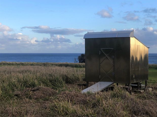 A small dark corrugated-metal shed or cabin-like structure stands in a coastal grassland, with a simple pitched metal roof, a central cross-braced door, and a narrow access ramp. The ocean and horizon are visible directly behind it under a partly cloudy blue sky. The image has a rugged, utilitarian coastal setting and subdued natural lighting.