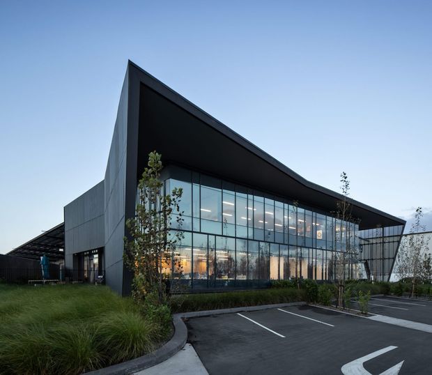Exterior view of a contemporary commercial building with a dramatic angular roof canopy, dark metal-clad side walls, and extensive floor-to-ceiling glass curtain walls. The building fronts a landscaped parking area with young trees and ornamental grasses, and warm interior lighting is visible through the glazing.