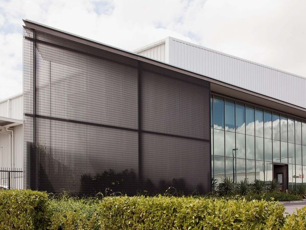 Exterior view of a contemporary commercial or institutional building with a long, low profile, white metal-clad upper volume, dark gridded screening across the facade, and a large glazed frontage reflecting the sky. Landscaped hedging sits along the foreground.