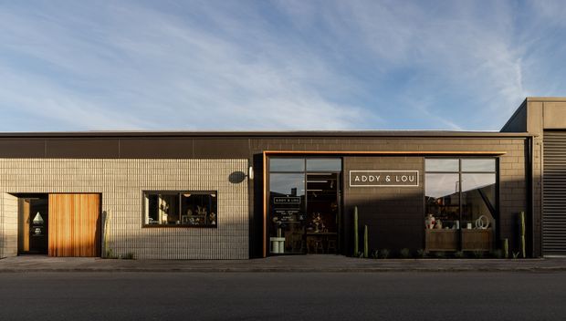 Street-facing contemporary commercial storefront with a low, rectilinear facade clad in small beige tiles and dark metal framing. Large glazed shopfront windows display the interior, with a prominent ADDY & LOU sign above the entrance. A timber slatted door and dark louvred facade element add material contrast.