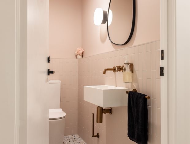 Compact, softly lit bathroom with a wall-mounted white basin, exposed brass-toned tap and plumbing, white toilet, patterned terrazzo-style floor, and pale blush-pink square wall tiles with matching painted upper walls. A rounded black-framed mirror and globe wall light add contemporary contrast.