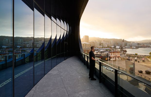 A sweeping curved balcony or terrace on a contemporary urban building, enclosed by floor-to-ceiling dark glass and framed by black structural elements. The broad grey tiled floor and glass balustrade overlook a dense waterfront cityscape and harbour at sunset. The dramatic curved facade is strongly reflected in the glazing, creating a sculptural, high-impact architectural composition.