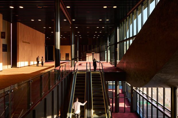 A spacious, multi-level contemporary interior atrium with escalators, glass balustrades, tall floor-to-ceiling glazing, dark slatted ceilings, and warm timber wall finishes. Strong sunlight enters from the right, illuminating a red-toned carpeted circulation area and open public lobby. The space appears institutional or commercial, with multiple people using the escalators and walkways.