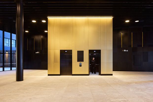 A spacious, high-end commercial lobby or lift foyer with a central gold-toned elevator core, dark wall finishes, black ceiling with recessed linear detailing, large pale stone floor tiles, and a cylindrical structural column. Floor-to-ceiling glazing on the left provides daylight, while illuminated signage and elevator doors reinforce the commercial building setting.