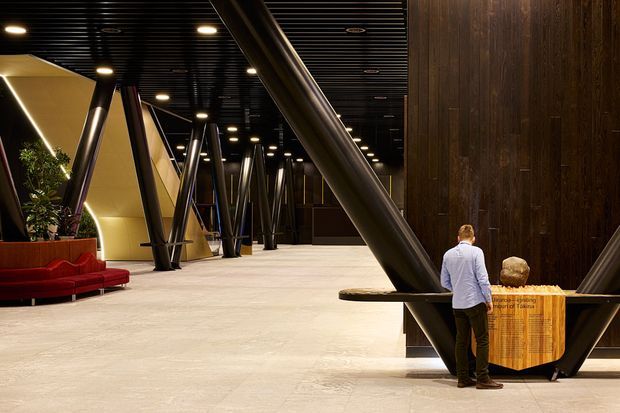 A dramatic contemporary commercial interior lobby or reception space with a broad open floor, black slatted ceiling, extensive dark timber wall cladding, and prominent angled structural columns. Warm recessed lighting illuminates sculptural cream-coloured wall partitions, a red upholstered seating area, indoor planting, and a reception or display counter in the foreground.