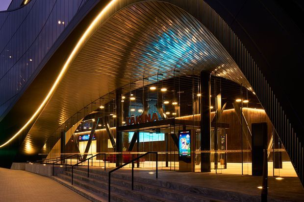 Night-time exterior view of a dramatic contemporary commercial building entrance. A sweeping curved canopy with ribbed metallic soffit and integrated linear lighting frames glazed entry doors, illuminated signage, digital displays and broad accessibility steps with black handrails. The architecture uses strong flowing geometry, dark cladding and warm interior lighting, creating a high-impact urban frontage.