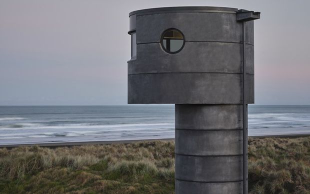Exterior view of a sculptural, cylindrical dark concrete structure positioned in coastal dunes, with the ocean and beach visible behind it. The structure has horizontal formwork bands, a small circular window, and a projecting flat roof or canopy, creating a stark minimalist architectural presence.