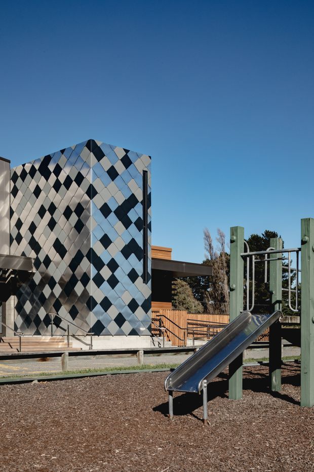 Exterior view of a contemporary public or educational building beside a playground. The building features a striking geometric diamond-patterned facade in dark blue, pale blue, silver, and cream panels, with timber-toned elements and accessible stairs and ramps. A metal playground slide and green-painted play structure occupy the foreground on a wood-chip surface under a clear blue sky.