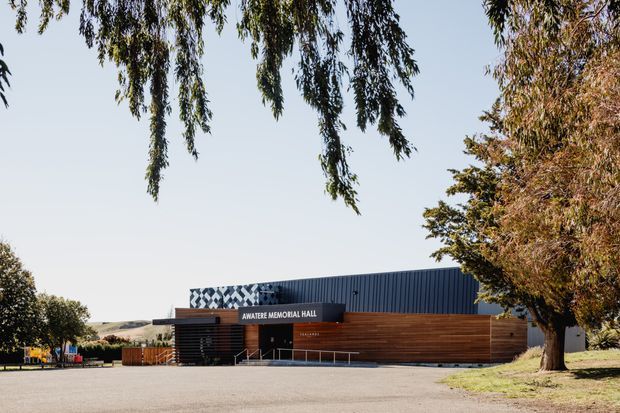 Exterior view of the Awatere Memorial Hall, a low-rise public/community building with a dark corrugated metal upper volume and warm horizontal timber-clad facade. The entrance canopy and accessible ramp are visible, with mature trees and open grounds surrounding the paved forecourt. A distinctive patterned geometric feature wall appears behind the roofline.