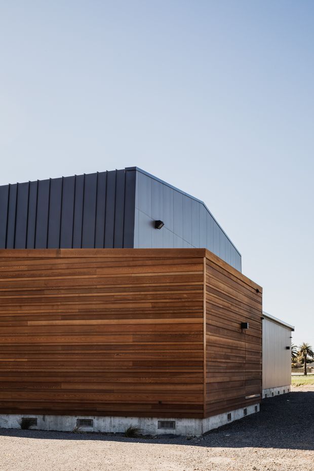Exterior view of a contemporary, rectilinear building with a warm horizontal timber-clad lower volume and a dark grey-blue standing-seam metal upper volume with a sloped roofline. The structure sits on a concrete base above a gravel surface, with minimal exterior lighting and a clear sky.