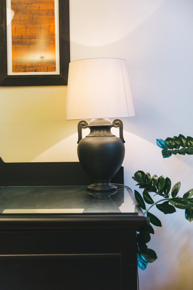 Close-up interior vignette featuring a traditional dark wood console or sideboard with a glossy top, an ornate black urn-shaped table lamp with a white fabric shade, framed abstract artwork, and leafy indoor plants. Warm lamp light creates a refined, softly layered atmosphere against pale walls.
