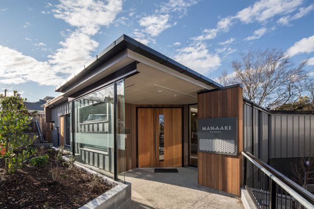 Contemporary low-profile building entrance with broad roof overhangs, vertical timber entry doors, dark metal cladding, extensive glazing, and a prominent timber sign panel reading “MANAAKI.” A paved walkway and landscaped planting lead to the sheltered entry, with a bright blue sky and mature trees in the background.
