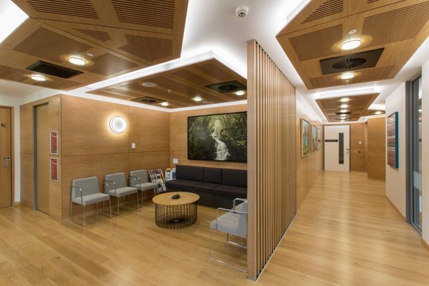 A contemporary commercial waiting area and corridor with warm timber finishes. The space features light wood flooring and wall panelling, a slatted timber acoustic ceiling and vertical timber screen, recessed lighting, a dark upholstered sofa, grey lounge chairs, a round coffee table, framed artwork and glazed office or room fronts along the hallway.