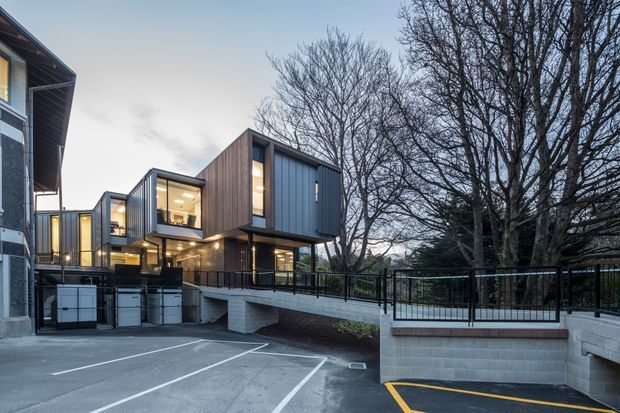 Exterior view of a contemporary multi-level building composed of offset box-like volumes, with dark vertical metal cladding, warm timber-clad sections, extensive glazing, and a raised terrace or walkway with black metal balustrades. The building is set beside a paved parking area and mature deciduous trees, photographed in cool evening light.