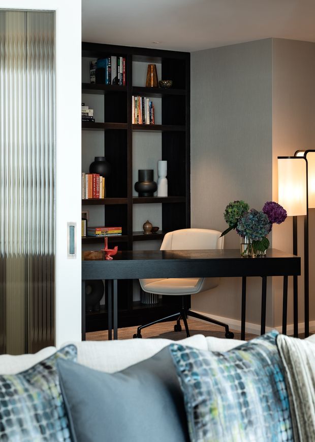 A refined home office viewed from a bedroom, featuring a black built-in bookshelf filled with books and decorative objects, a dark timber desk, a light upholstered office chair, and a contemporary floor lamp. A partially open ribbed-glass sliding door and the foreground bed with layered blue and patterned cushions are visible. The room has a sophisticated, moody palette with warm ambient lighting.