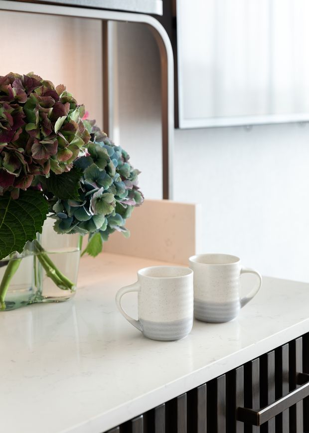 Close-up interior view of a bright kitchen or dining counter, featuring a white marble-look benchtop with two ceramic mugs and a glass vase of hydrangeas. Dark vertical slatted cabinetry or panelling sits beneath the counter, with warm timber detailing and a large light-coloured window or wall panel in the background.
