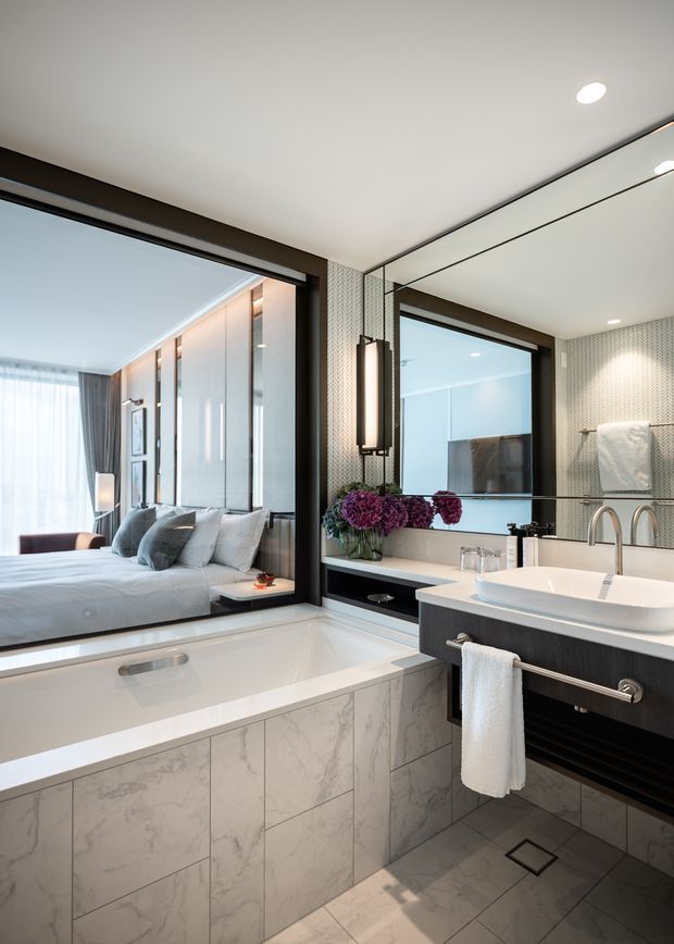 A refined modern bathroom viewed toward a bedroom through a large internal glazed opening. The room features a freestanding-style white bathtub set into a marble-look tiled surround, a wall-mounted dark vanity with a white vessel basin, large mirror, chrome tapware, patterned wall tiles, integrated lighting, and white towels. The adjacent bedroom is visible through the glazing and reflection.