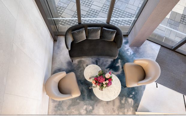 A contemporary lounge seating area viewed from above, with a curved dark sofa, two rounded armchairs, and a small round pale stone coffee table arranged on a large abstract blue-grey rug. Floor-to-ceiling glazing and a patterned paved exterior are visible beyond the seating, with a tall light-coloured wall and structural column framing the space.