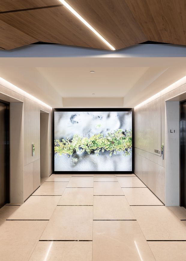 A refined modern elevator lobby or hallway with pale stone tiled flooring and large light-toned wall panels. Recessed linear lighting runs along the ceiling edges, while a warm timber-lined angled ceiling feature frames the upper portion of the space. A large illuminated botanical artwork forms a focal point at the end, with elevator doors visible on both sides.