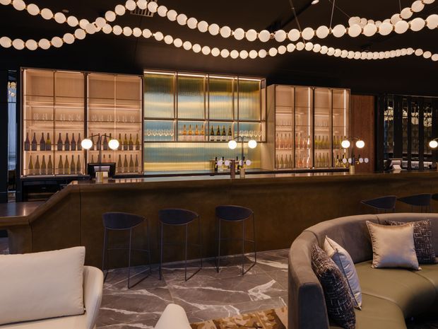 A sophisticated, dimly lit cocktail bar or lounge with a long dark bar counter, illuminated glass-backed bottle displays, high stools, and curved upholstered seating in the foreground. Decorative strings of glowing round lights span the dark ceiling, while the floor features large marble-look slabs with strong veining. The atmosphere is luxurious, intimate, and dramatic.