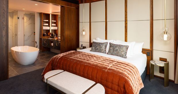 A sophisticated bedroom with a freestanding oval bathtub visible through an open ensuite area. The room features a large upholstered bed, a deep rust-brown quilted throw, patterned accent cushions, a padded bench, dark timber wall panels, cream wall panels with timber trim, and pendant bedside lights. Dark stone or tile flooring extends into the ensuite, which includes a freestanding bath and illuminated built-in shelving.
