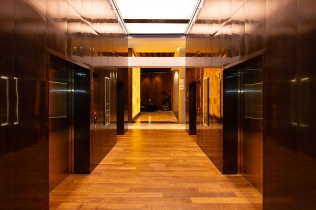 A symmetrical, richly finished elevator lobby or hallway with dark glossy wood wall panels, multiple black elevator doors, warm timber flooring, and a bright recessed ceiling light. The reflective surfaces and amber lighting create a polished, luxurious atmosphere.