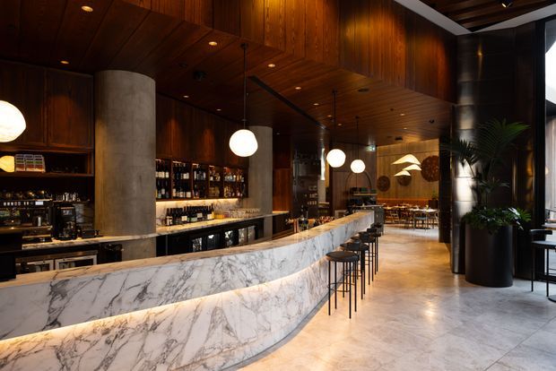 A sophisticated hospitality-style bar and dining space with a long curved marble bar, dark timber wall and ceiling panelling, integrated bottle shelving, black bar stools, globe pendant lights, and a concrete cylindrical column. The warm, dimly lit interior extends toward a dining area and includes large potted greenery near the glazing.