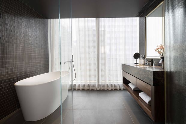 A contemporary bathroom with a freestanding white bathtub beside a clear glass shower enclosure. Dark textured mosaic wall tiles contrast with large grey floor tiles, while a timber vanity with a dark stone benchtop, integrated basin, round mirror, and open towel storage sits beneath a large window with sheer curtains. The room has a refined, spa-like atmosphere and abundant diffused natural light.