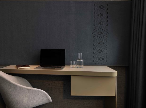 A compact, moody home workspace with a floating light-toned desk against a dark textured feature wall. A laptop, notebook and glassware sit on the desk, accompanied by a softly upholstered chair. Dark curtains frame the right side, while a vertical geometric patterned wall detail adds visual interest.