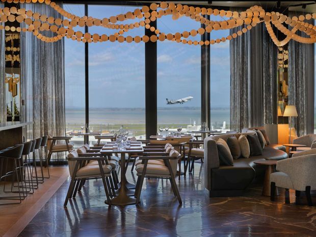 A stylish waterfront restaurant or dining lounge with floor-to-ceiling glazing overlooking the beach and ocean. The interior features numerous dining tables and chairs, a long upholstered banquette, lounge seating, bar stools, dark reflective flooring, sheer curtains, warm pendant and table lighting, and decorative garlands suspended across the ceiling. A plane is visible outside over the waterfront.