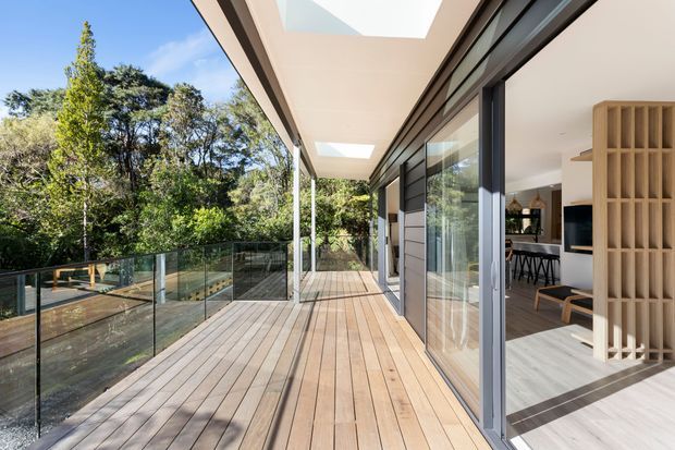 A spacious timber deck terrace runs alongside a contemporary home, bordered by clear glass balustrades and surrounded by mature greenery. Full-height glazed sliding doors open into a bright interior with a kitchen or dining area, dark-framed windows, pale flooring, and a vertical timber-screen partition. A broad soffit with multiple skylights shelters the deck.