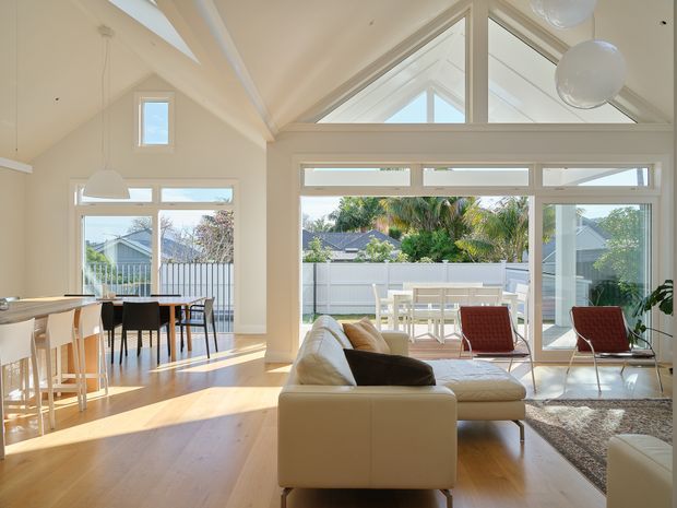 Bright, airy open-plan living and dining area with high pitched ceilings, extensive white-framed glazing, and a glazed triangular clerestory. A pale timber floor, cream sectional sofa, dining table with mixed seating, and contemporary pendant lights create a warm modern interior. The glazing opens toward a furnished outdoor terrace and landscaped suburban surroundings.