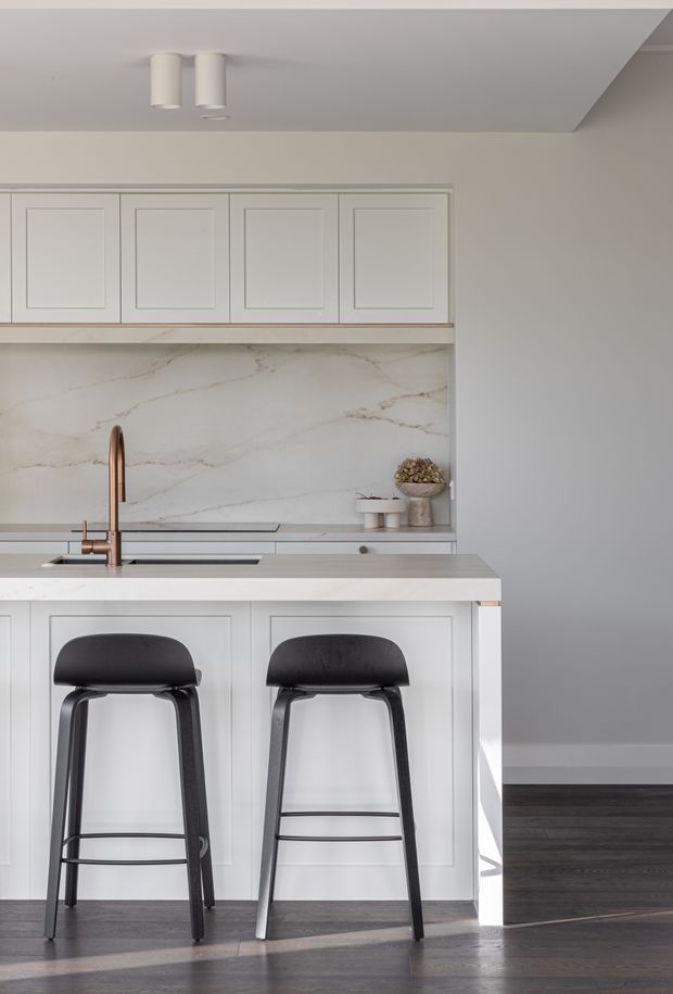 A bright, refined kitchen with white shaker-style cabinetry, a large veined stone backsplash and benchtop, an integrated island with two black bar stools, and a bronze-toned curved faucet. Dark timber flooring contrasts with the predominantly white and light-grey fit-out. Recessed and cylindrical ceiling lights provide simple illumination.