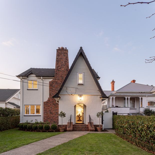 Front exterior view of a compact period-style house with a steep front gable, painted weatherboard cladding, exposed brick chimney, leadlight windows, covered entry, and a central paved path through a manicured garden. A neighbouring traditional timber house is visible to the right.
