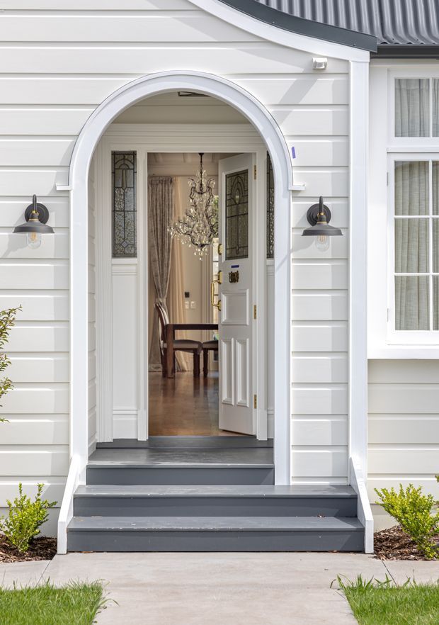 Front exterior view of a refined white weatherboard home entrance. The facade features a tall arched portico opening with layered white trim, dark grey timber steps, traditional wall-mounted lanterns, and a partially open glazed panelled front door. A chandelier, drapery, and dining furniture are visible inside the entry, while a multi-pane window and corrugated metal roof appear to the right and above.