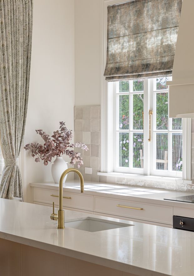 Bright, refined kitchen interior featuring a large white stone island with an undermount sink and brushed brass gooseneck tap. Pale cabinetry with brass bar handles runs beneath a sunlit multi-pane window, with a matching pale stone benchtop and a subtle tiled splashback. Patterned floor-to-ceiling drapery and a textured Roman blind frame the window, while a vase of dried foliage adds a muted organic accent.