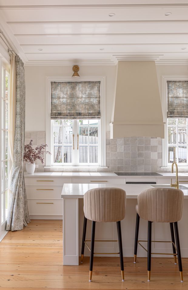 Bright, refined kitchen with a large island and two upholstered bar stools. The room features white shaker-style cabinetry with brass pulls, a pale stone benchtop, a grey-beige tiled splashback, a sculptural rangehood surround, timber flooring, tall sash-style windows with patterned Roman blinds and curtains, and an ornate coffered or panelled ceiling.