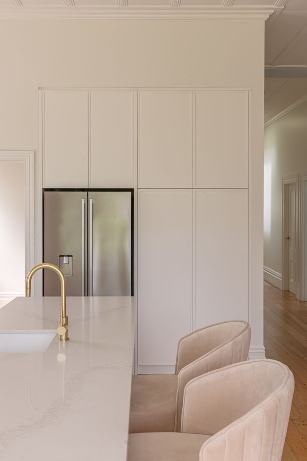 A refined, light-toned kitchen centred on a large pale stone island with a brushed brass gooseneck tap and integrated seating. The wall behind features full-height, recessed-panel cabinetry concealing storage and framing a stainless-steel double-door refrigerator. Upholstered neutral bar stools line the island. Decorative ceiling cornices, wall mouldings, and the adjoining timber-floored hallway create a classic, elegant character.